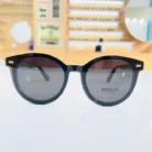 Gros plan sur lunettes rondes noires, monture épaisse brillante, verres gris et détails métalliques aux coins.