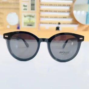 Gros plan sur lunettes rondes noires, monture épaisse brillante, verres gris et détails métalliques aux coins.