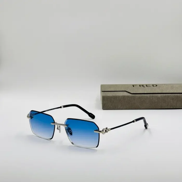 Lunette de soleil Fred métal verres bleus dégradés Lunette de soleil Fred en métal avec verres bleus dégradés, branches fines à embouts noirs, posée près d’un étui gris FRED.