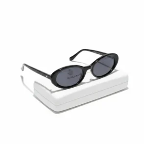 Lunette de soleil femme noire à forme ovale, verres teintés foncés, tenue à la main au-dessus d’un étui blanc.