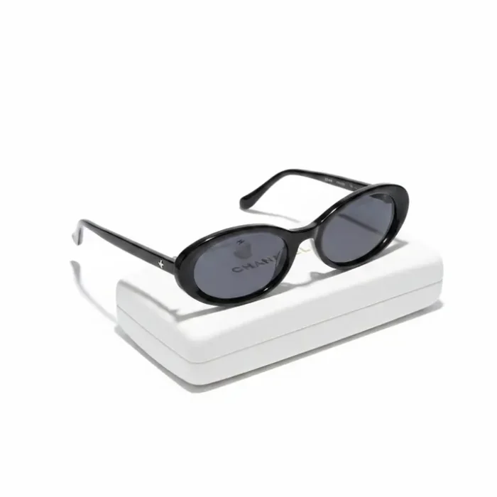 Lunette de soleil femme noire ovale avec étui Lunette de soleil femme noire à forme ovale, verres teintés foncés, tenue à la main au-dessus d’un étui blanc.